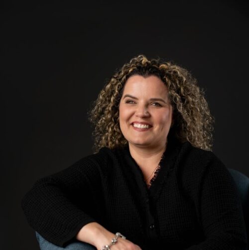 Foto de la coach Johanna Alvarado, sentada con blusa color negro, sonriendo con cabello rubio en colochos y las manos cruzadas al frente.