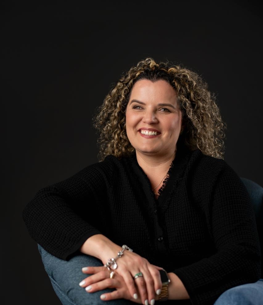 Foto de la coach Johanna Alvarado, sentada con blusa color negro, sonriendo con cabello rubio en colochos y las manos cruzadas al frente.